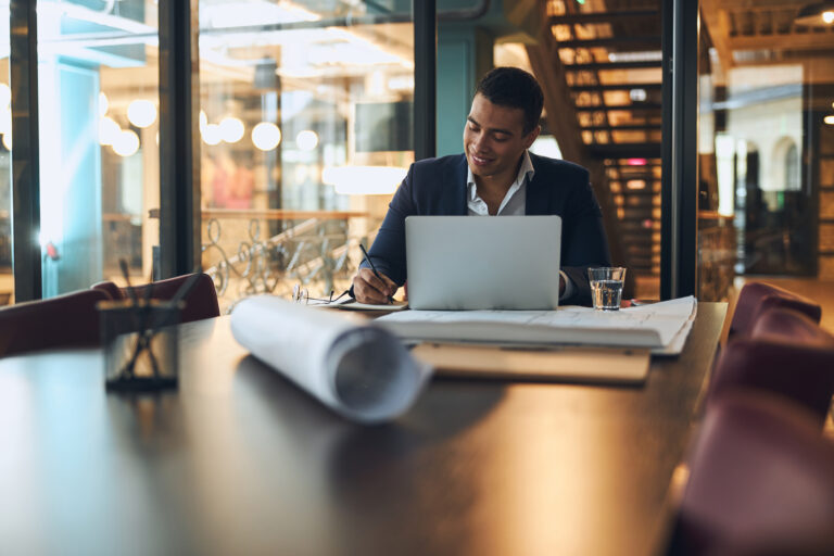 Henkilö istuu pöydän ääressä katsoo kannettavaa tietokonetta. Pöydällä on lasi ja papereita.